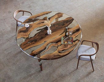 May include: An overhead view of a round dining table with a unique wood grain design. The table is surrounded by two chairs with light gray cushions. A vase, wine bottle, and book are on the table.