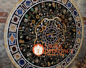 May include: A round black marble table top with intricate floral and geometric inlay designs. The table features a border of alternating shapes and colors, with a central floral medallion. The text "AGRA HERITAGE Marble Crafts" is visible.