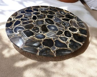 May include: A round table top made of agate slices. The agate is a mix of black, gray, and brown colors. The table top is polished and smooth.