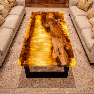 May include: A rectangular coffee table with a unique design. The tabletop features a translucent, amber-colored resin with embedded wood grain. The table is supported by a black, rectangular metal frame. The table is surrounded by beige sofas.