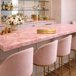 May include: A luxurious home bar with a pink marble countertop and matching pink velvet bar stools with gold legs. The bar features a mirrored back with shelves displaying glassware and bottles. A floral arrangement adds a touch of elegance.