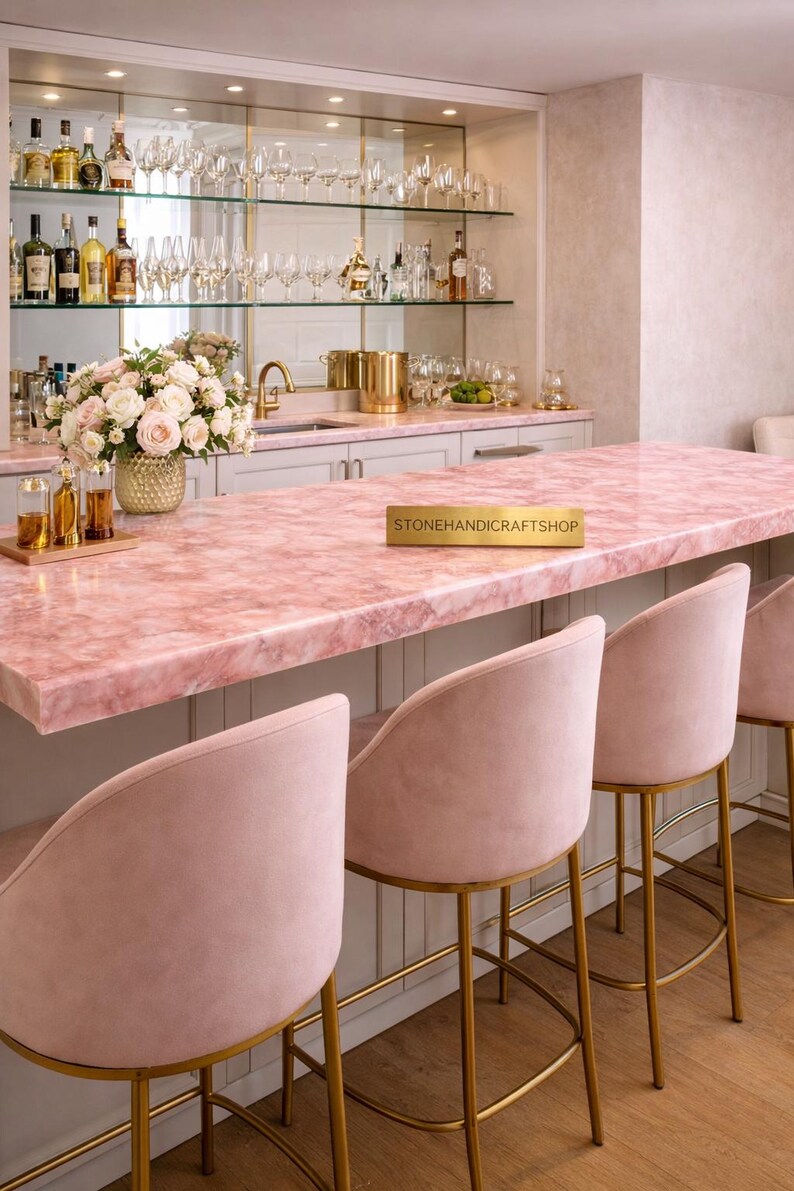 May include: A luxurious home bar with a pink marble countertop and matching pink velvet bar stools with gold legs. The bar features a mirrored back with shelves displaying glassware and bottles. A floral arrangement adds a touch of elegance.