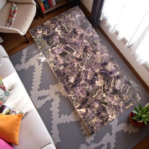 May include: A rectangular amethyst coffee table with a polished surface. The table's surface is a mosaic of deep purple, gray, and white crystalline fragments. The table is set on a gray patterned rug, with a sofa and chair visible.