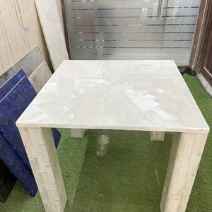 May include: A white square table with a white marble top and four white marble legs. The table has a unique design with a circular pattern on the top.