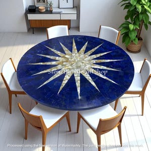 Puede incluir: Una mesa de comedor redonda azul con un diseño de estrella en el centro. Seis sillas blancas con marcos de madera rodean la mesa. La superficie de la mesa es un mosaico de piedras azules y blancas. El texto "StoneHandicraftShop" es visible.