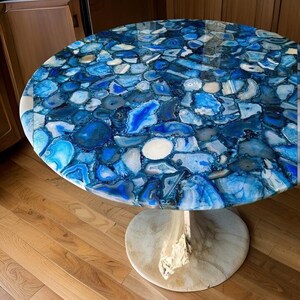 Könnte beinhalten: Ein runder Tisch mit einer blauen Achatplatte und einem weißen Sockel. Die Tischplatte besteht aus vielen kleinen Achatstücken, jedes mit einem einzigartigen Muster. Der Sockel ist ein schlichtes, elegantes Design.