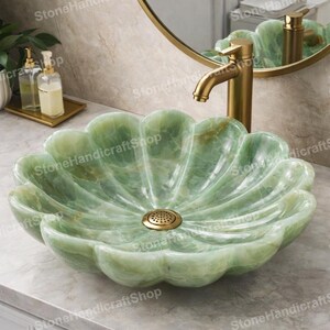 May include: A green onyx stone sink with a flower petal design. The sink has a gold-colored drain and is paired with a gold faucet. The sink is set against a neutral-toned wall and a gold-framed mirror.