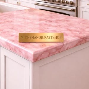 May include: A pink marble countertop with a veined pattern. The countertop is installed on white cabinets. A gold rectangular sign with the text "STONEHANDICRAFTSHOP" is on the countertop.