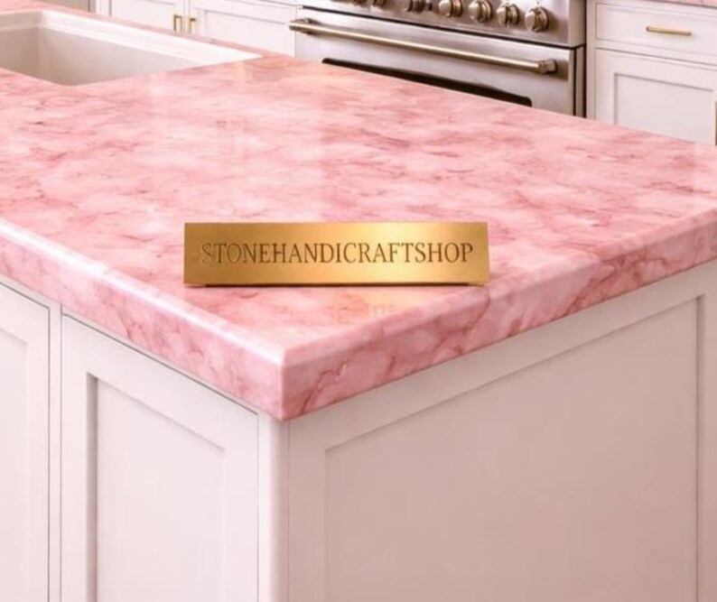 May include: A pink marble countertop with a veined pattern. The countertop is installed on white cabinets. A gold rectangular sign with the text "STONEHANDICRAFTSHOP" is on the countertop.