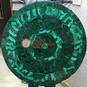 Puede incluir: Una mesa redonda de malaquita verde con un patrón de mosaico. La superficie presenta una mezcla de tonos verdes oscuros y claros. La mesa presenta el texto "AGRA HERITAGE Marble Crafts".