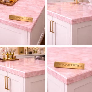 May include: Pink marble countertop with white cabinets and gold hardware. The countertop has a polished finish and is accented by a wooden tray with bottles and a sign that reads "STONEHANDICRAFTSHOP".