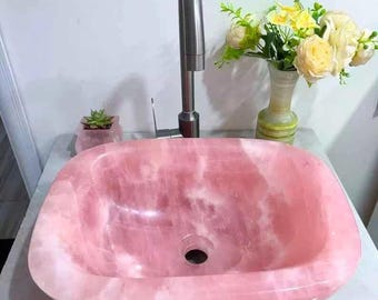 May include: A pink, oval-shaped stone sink with a polished finish. A stainless steel faucet is mounted above the sink. A small succulent plant in a pink pot and a vase of flowers are on the countertop.