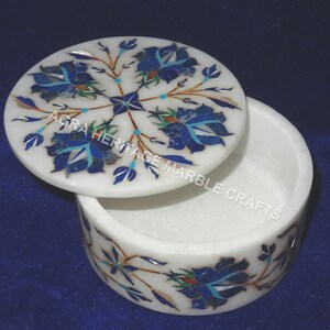 May include: White marble trinket box with a lid featuring a floral design in blue and teal. The box is round with a matching floral pattern around the base. The text "AGRA HERITAGE MARBLE CRAFTS" is printed on the side of the box.