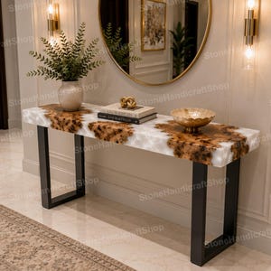May include: A rectangular console table featuring a white and brown natural stone top with organic patterns, supported by two black metal U-shaped legs. The surface holds a textured vase with greenery, two stacked books, and a gold-toned bowl.