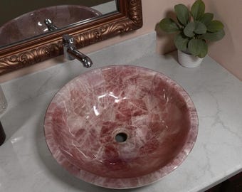 Pode incluir: Uma pia redonda de pedra rosa com um padrão semelhante a um mosaico. A pia está colocada sobre uma bancada branca, com uma torneira cromada e um espelho decorativo ao fundo. Uma pequena planta verde num vaso branco também é visível.