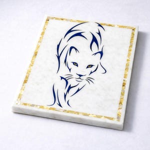 Puede incluir: Posavasos cuadrado de mármol blanco con un diseño de pantera azul. La pantera tiene ojos amarillos y está delineada en azul. El posavasos tiene un borde dorado hecho de pequeños trozos irregulares. Un artículo decorativo para el hogar.