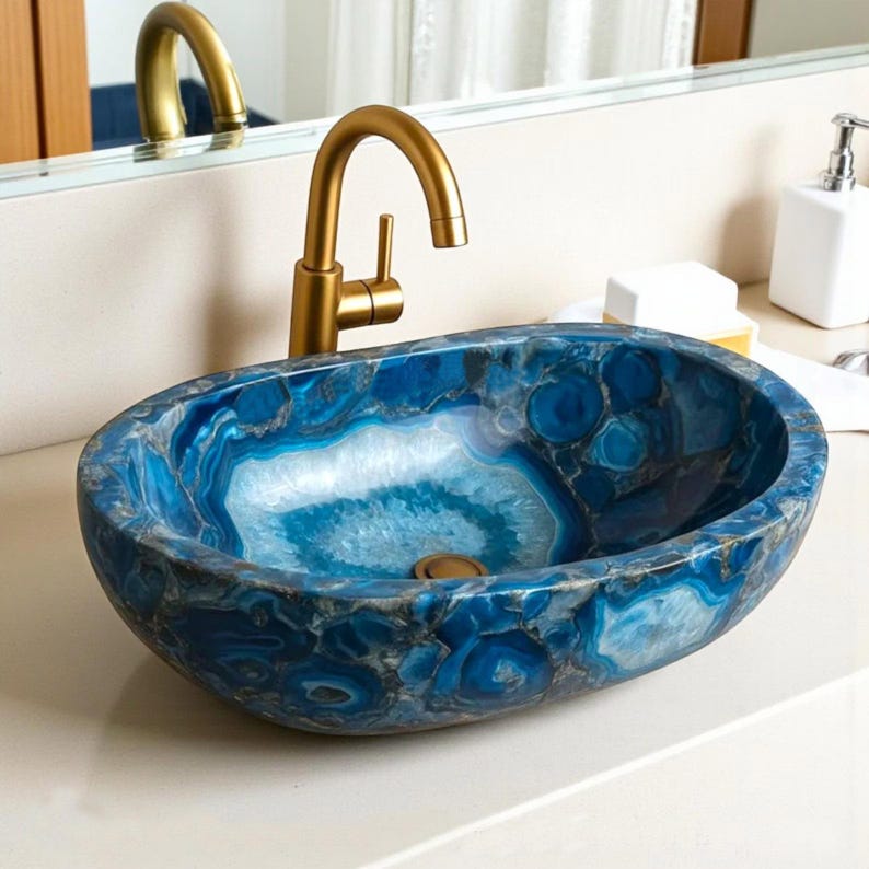 May include: Oval-shaped blue agate stone sink with a gold-colored faucet. The sink has a unique pattern of blue and white swirls and circles. The faucet is a gooseneck style with a single handle. The sink is set on a white countertop.