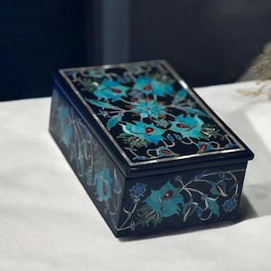 May include: A black wooden box with a floral design. The box is decorated with blue, green, and white flowers and leaves. The box has a silver trim around the edges.