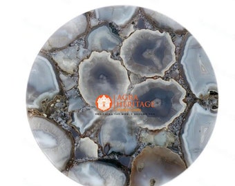May include: A round, polished stone tabletop with a mosaic pattern of gray, white, and brown agate slices. The agate pieces are arranged in a circular design, creating a unique and natural look. The tabletop is suitable for a small table or decorative display.