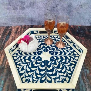 May include: A hexagonal serving tray with a blue and white floral design. The tray has a bone inlay design and is made of wood. Two wine glasses are on the tray, along with a white decorative object.