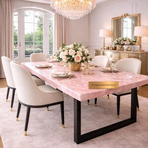May include: A dining room set featuring a pink quartz table with black legs, surrounded by cream-colored chairs. A gold chandelier hangs above the table, which is set with plates and glasses. A floral arrangement sits in the center.