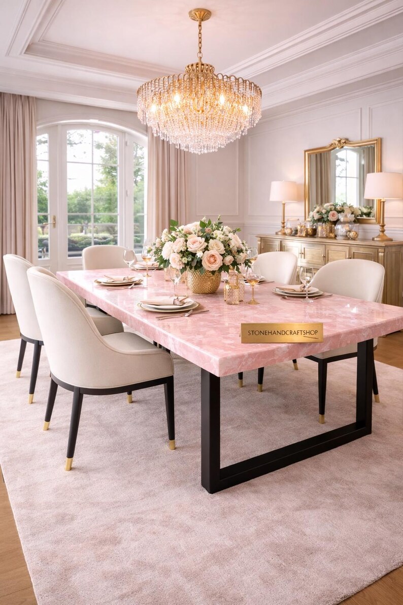 May include: A dining room set featuring a pink quartz table with black legs, surrounded by cream-colored chairs. A gold chandelier hangs above the table, which is set with plates and glasses. A floral arrangement sits in the center.