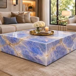 May include: A large, square coffee table with a blue and gold marble design. The table is the focal point of a living room, with a sofa, armchair, and rug. A gold tray with flowers and a candle sits on the table.