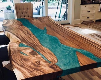 Puede incluir: Una mesa de comedor rectangular con un tablero de madera de borde natural que presenta una incrustación central fluida de resina turquesa translúcida. La madera muestra un veteado marrón intenso y se apoya sobre una base de metal oscuro.