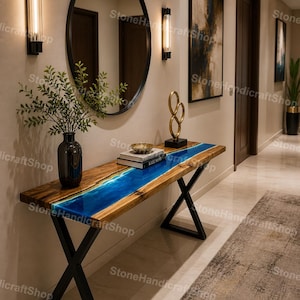 Op de afbeelding: Een houten console met een blauw hars rivierontwerp. De tafel heeft een donkere houten afwerking, zwarte metalen poten en is versierd met boeken en een gouden sculptuur. De muur achter de tafel heeft een spiegel en wandkandelaars.
