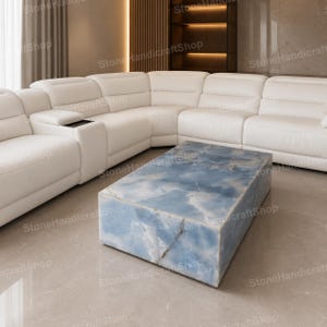 Könnte beinhalten: Ein rechteckiger Couchtisch aus blauem Onyx-Stein mit weißer Maserung, der eine glatte, polierte Oberfläche und eine massive Blockkonstruktion aufweist. Der Tisch steht vor einem großen weißen Ledersofa mit integrierten Sitzgelegenheiten.
