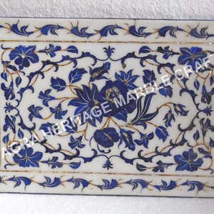 Puede incluir: Una bandeja de mármol blanco con un diseño floral azul y dorado. La bandeja está incrustada con lapislázuli y madreperla. El texto "AGRA HERITAGE MARBLE CRAFTS" es visible en el centro de la bandeja.