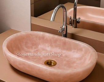 May include: Oval-shaped pink stone sink with a polished brass drain and a chrome faucet. The sink has a smooth, marbled appearance and is set against a neutral background. The text "StoneHandicraftShop" is visible on the sink.