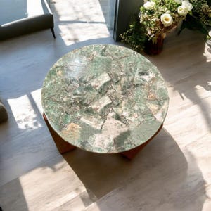 May include: A round coffee table with a green and white stone top. The table has a wooden base and is set on a light-colored wooden floor. The table's top is made of a mosaic of green and white stones.