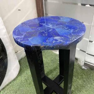 Op de afbeelding: Een kleine ronde bijzettafel met een blauw lapis lazuli stenen blad en een zwarte metalen onderstel. Het onderstel is een eenvoudig kruis.