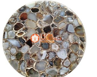 May include: A round, decorative tabletop made of polished agate stones in shades of gray, brown, and white. The stones are arranged in a mosaic pattern, with gold-colored edges. The tabletop has a glossy finish and is set against a white background.