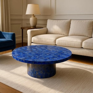 May include: A round coffee table with a mosaic-style top in shades of blue. The table has a cylindrical base and sits on a light-colored rug. A beige sofa and blue armchair are in the background.