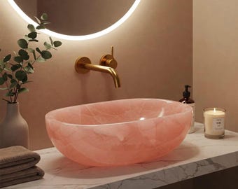 May include: Oval-shaped pink stone bathroom sink. The sink is set on a marble countertop with a gold faucet and a round mirror in the background. A lit candle and a bottle of soap are also visible.
