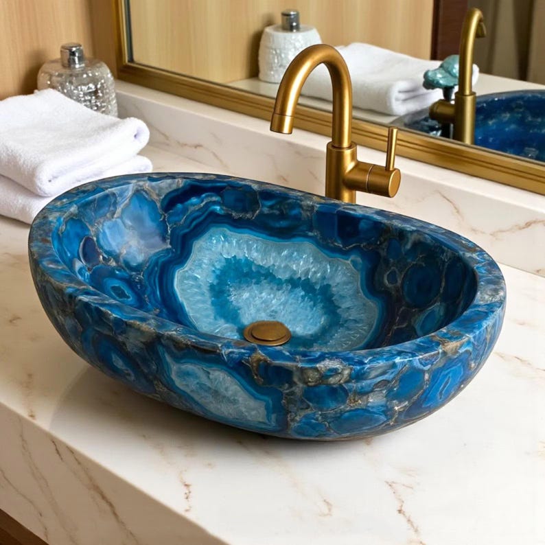 May include: An oval-shaped, blue agate stone sink with a gold faucet. The sink has a unique pattern of light and dark blue hues, with a central lighter blue design. The faucet is a curved, single-handle design. The sink is set on a white countertop.