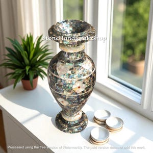 May include: A decorative mosaic vase with a wide top and a flared base. The vase is adorned with iridescent tiles and inlaid with white flower designs. The vase is approximately 12 inches tall and sits on a white surface near a window.