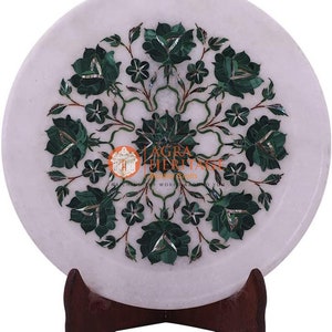 Op de afbeelding: Witte marmeren plaat met een bloemeninlegontwerp. Het ingewikkelde patroon toont groene malachietbloemen en bladeren in een cirkelvormig patroon. De plaat wordt gepresenteerd op een donkerbruine houten standaard, wat het vakmanschap benadrukt.