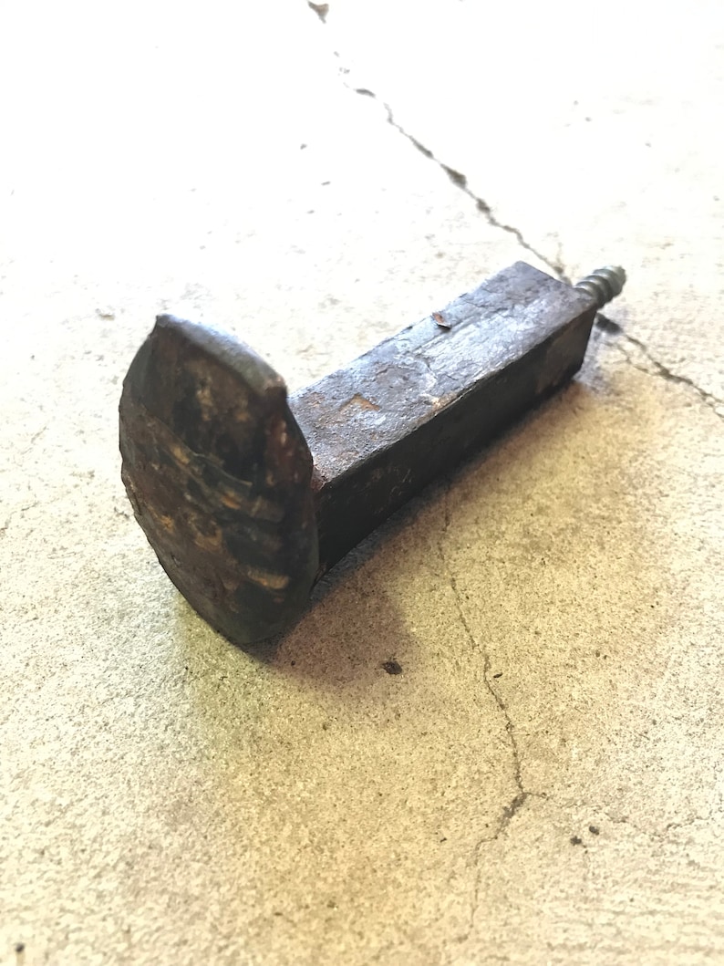 Op de afbeelding: Een roestige metalen spijker met een vierkante kop en een schroefdraad aan het uiteinde. De spijker ligt op een betonnen oppervlak.