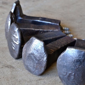 Op de afbeelding: Vijf grote, zware, metalen spoorwegspijkers met zeskantige koppen. De spijkers zijn in een halve cirkel gerangschikt met de koppen naar de kijker gericht. De spijkers zijn donkergrijs van kleur en hebben een ruw, getextureerd oppervlak.