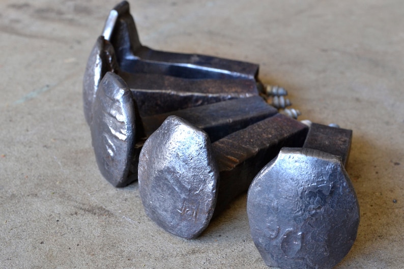 Op de afbeelding: Vijf grote, zware, metalen spoorwegspijkers met zeskantige koppen. De spijkers zijn in een halve cirkel gerangschikt met de koppen naar de kijker gericht. De spijkers zijn donkergrijs van kleur en hebben een ruw, getextureerd oppervlak.