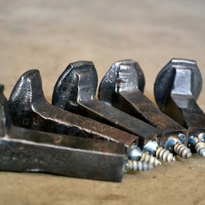 Op de afbeelding: Vijf rustieke metalen haken gemaakt van hergebruikte spoorwegspikes. Elke haak heeft een schroef aan de onderkant voor eenvoudige installatie. De haken zijn donkerbruin van kleur met een ruw, getextureerd oppervlak.