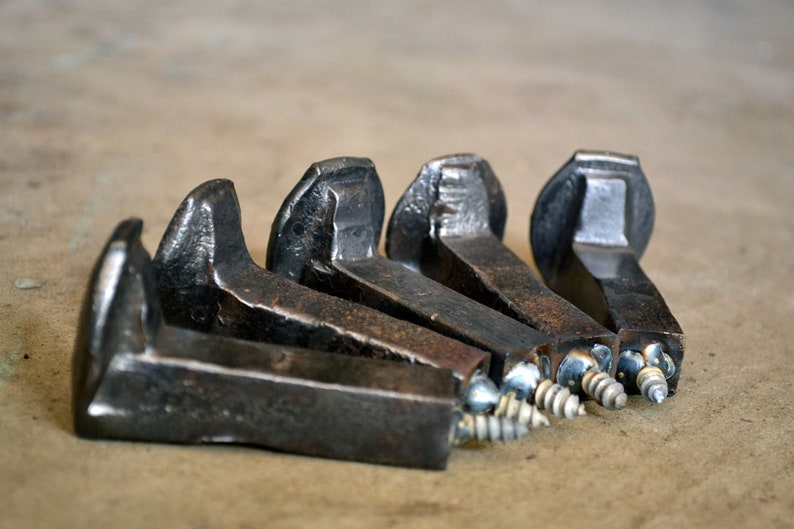 Op de afbeelding: Vijf rustieke metalen haken gemaakt van hergebruikte spoorwegspikes. Elke haak heeft een schroef aan de onderkant voor eenvoudige installatie. De haken zijn donkerbruin van kleur met een ruw, getextureerd oppervlak.