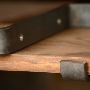 Puede incluir: Primer plano de un soporte de estante de metal negro con un borde curvo. El soporte está montado en un estante de madera.