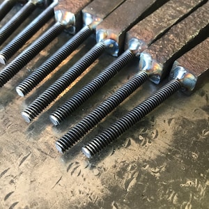 Op de afbeelding: Close-up van een groep metalen staven met schroefdraad aan de uiteinden. De staven zijn in een rij gerangschikt en zijn allemaal even groot. De uiteinden van de staven zijn vierkant gelast.