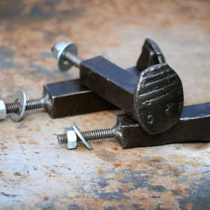 Op de afbeelding: Drie zwarte metalen spoorwegspijkerhaken met zilveren moeren en ringen. De haken zijn bevestigd aan een vierkante metalen basis.