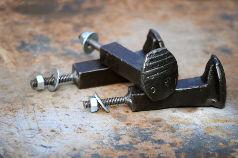 Op de afbeelding: Drie zwarte metalen spoorwegspijkerhaken met zilveren moeren en ringen. De haken zijn bevestigd aan een vierkante metalen basis.