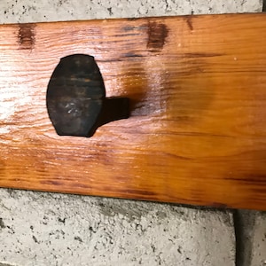 Op de afbeelding: Een rustieke houten wandhaak met een enkele, grote, zwarte metalen haak. De haak is bevestigd aan een houten plank die is gemonteerd op een witte bakstenen muur.